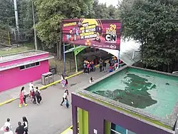 Several individual and groups of people in an adventure park, with a signboard of an event's advertisement and surrounded with the dense trees under a sunny sky
