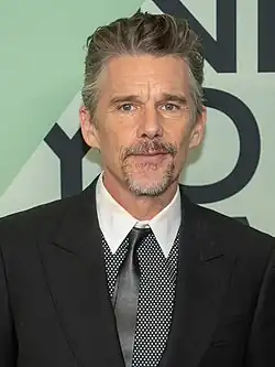 A Caucasian male with gray hair, wearing a two-piece dark gray suit with a gray and white shirt.