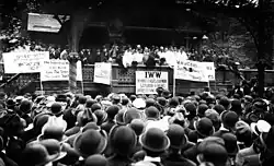 Black and white photograph of a speaker rallying a large crowd. In front of the stage, facing the audience, are several signs, in various languages, displaying demands.