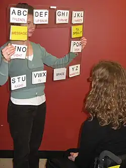 a user in a wheelchair faces a communication partner who holds a transparent piece of plastic between them, the plastic has the letters of the alphabet printed around the edges