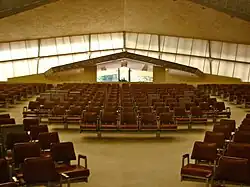 Westward view of the main sanctuary, looking toward the main entrance. There are seats in the foreground. The windows on the upper part of the base are visible in the background; these windows are slanted upward, and the entrance is visible beneath the peak of the windows.