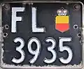 Two-line Liechtenstein license plate in the then-standard square format of 310 × 240 mm, with the gold and red escutcheon of the Prince's coat of arms as a sticker, instead of the painted national coat of arms. These plates were issued between 1961 and 1972.