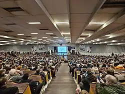 Wide angle photograph of the nearly full Jansen auditorium at ULB from the upper stairs looking down. Depicting people queueing on the sides to find an empty seats while the speaker prepares to give the FOSDEM 2025 Closing Talk.