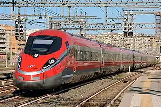 A red streamlined passenger train rolls through a station, with catenary wires above