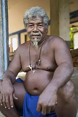 Elderly man in blue loincloth with keys on neck lace.