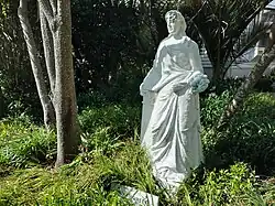A white stone statue of an adult woman wearing a long, elegant dress. She holds a bouquet that has been painted blue.