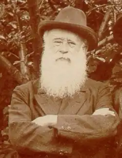 A sepia-tone photograph of a man from the chest up. He is wearing a hat and sports a long white beard. His arms are crossed.