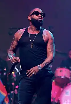 Ferré Gola wearing dark pants and a dark see-thru tank top, with black shades and crucifix necklace, standing onstage with microphone in right hand, band in background