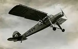 Black-and-white photo of a small propeller airplane, seen from below while climbing. Its landing gear is long and not foldable. It sports a Nazi Germany tail decoration and the sign D-IKVN.