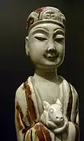 A grey statue of a thin figure in a flat topped hat and loose robes holding a piglet. The robes and hat are painted brown.