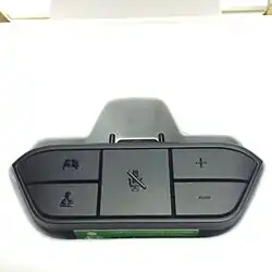 Xbox One Stereo Headset Adapter, a black plastic attachment that plugs into the bottom of the Xbox One Wireless Controller and provides a standard 3.5 mm stereo headset jack with microphone contact. The adapter itself is roughly trapezoidal and has five buttons on its face; two buttons on the left to balance audio output between in-game chat (bottom) and game (top); a large center button to mute the chat microphone; and two buttons on the right to increase (top) or decrease (bottom) overall volume.