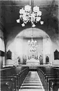 A view inside the Saint Mary Church in Lansing after combination gas-electric lighting was added. Note the balconies in the wings of the transepts, as well as the three arches in place of what had previously been load-bearing exterior walls. The plaster ceiling was decorated with a simple frieze and covered an attic or loft space.