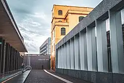 Under an overcast sky, a brick aisle runs between a brown building on the left, and a golden structure and array of glass windows split by grey columns on the right; in the near distance is a brown wall with white letters spelling out "FONDAZIONE PRADA". Another building, several stories tall, lies in the background.