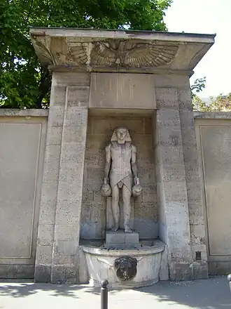 Stone fountain with statue of pharaoh holding jars, framed by wall with carved eagle above and lion head spout below