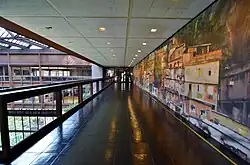 A footbridge on one of the upper stories. There is a handrail overlooking the atrium to the left, and a mural to the right.