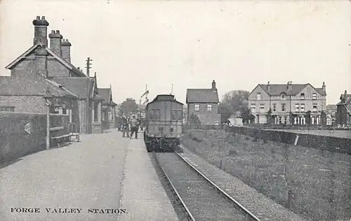 A black and white image of a train static in a station, with the platform on the left and houses beyond on the right