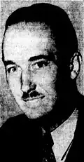 Black-and-white photographic portrait of a smiling man with slicked-back hair, a jagged nose and a divided toothbrush mustache. He wears a suit.