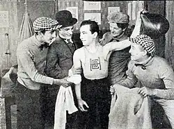 group of five men laugh and chat in a locker room, with one man wearing an athletic tank top as the others gesture and smile around him