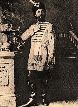 Monochrome photograph of a man standing, wearing leather books, knee-length tunic and feathered hat