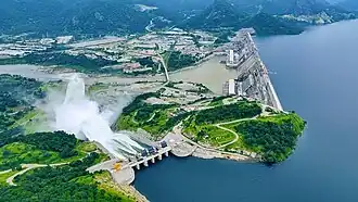 An photograph of large concrete dam and the reservoir it creates. The photo is taken from an aerial viewpoint, looking down from above. Large plumes of white water spray over a spillway.