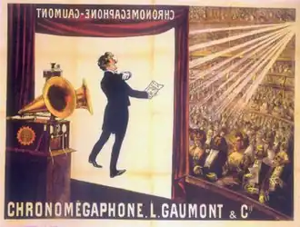 Illustration of a theater from the rear right of the stage. At the front of the stage, a screen hangs down with the projected image of a tuxedoed man holding up a text and performing. In the foreground is a gramophone with two horns. In the background, a large audience is seated at orchestra level and on several balconies. The words Chronomégaphone and Gaumont appear at both the bottom of the illustration and, in reverse, at the top of the projection screen.