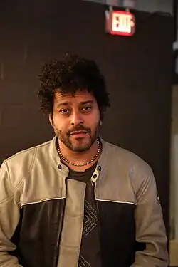 George William Lewis Jr., aka Twin Shadow, backstage wearing a brown print t-shirt under a leather jacket that is tan at the shoulders and dark brown under the arms, with silver bead necklace, looking directly at camera
