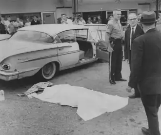 People stand around Rockwell with a sheet over his body