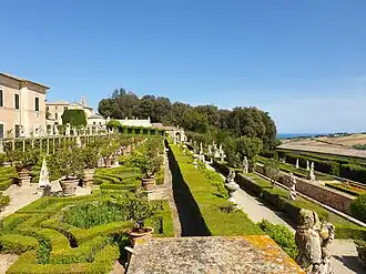 Terraced Italian-style garden with geometric hedges, stone statues, and a view over cultivated hills and the Adriatic Sea.