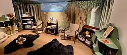 Small room with a rugs and children's books on small bookshelves. There is a painted background of a wooded prairie scene.