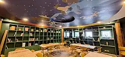 A small library room with desks, chairs, and books on shelves. The ceiling is painted with animals from the Illinois woodlands.