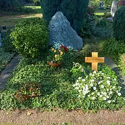A stone monument that is shaped like a mountain, alongside a wooden cross, surrounded by greenery