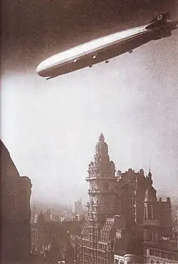 A sepia photograph of the airship from below. It is flying from right to left above a city. There is a tall ornate building just below the airship and the silhouette of more masonry in the bottom left foreground. All five engine nacelles are visible, and the two nearest and the control room are in sunlight. There are swastikas on both the vertical fins.
