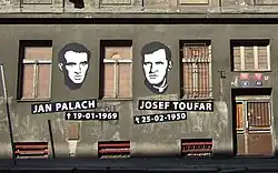 Graffiti between windows on the outside of a run down building, on the left ther is a painting of young mans face with text saying "JAN PALACH" then a cross symbol and the date "19-01-1969" and on the right there is a second face with text under it saying "JOSEF TOUFAR" also with a cross symbol and a date "25-02-1950".