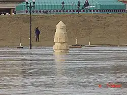 Water from a river completely surrounds a stone obelisk. In the background are several people with news cameras. A building with a green roof is seen behind them. At the bottom right of the image is the camera's time: "6 7:59&nbsp;am".
