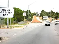 View of gavel-filled lane leaving the side of the road and then rising up a slope. Signs either side of it read 'TRUCK ARRESTOR BED'.