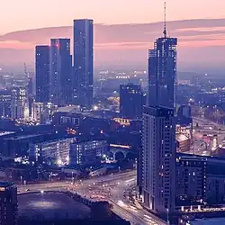 An aerial image of a city at night. There are five skyscrapers in the foreground and the photo is misty with tall hills and low-lying clouds in the background. The lights of the rest of the city can be seen in the background but not the buildings.