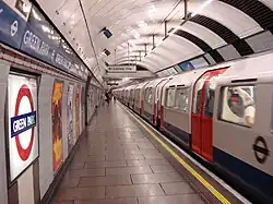 The interior of a building with a rounded, white roof, white walls, grey flooring, and a train on the right carrying passengers