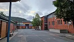 Three buildings with a parking lot in front of them and a tree-covered hill behind them