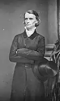A young man with long, combed-back hair, wears a dark cloak and holds a stovepipe hat