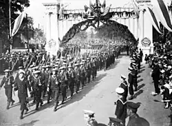 A group of men wearing military uniforms and carrying rifles marching under a decorated arch. The group is being led by a man carrying a sword. They are being watched by other men in uniform and a crowd of civilians.