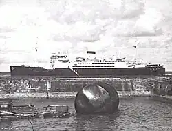Black and white photo of a large ship anchored off a beach. A damaged buoy is in the foreground of the photo.