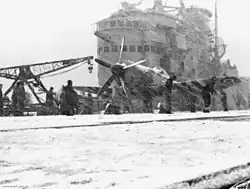 The flight deck of an aircraft carrier during a snowstorm, with the ship's superstructure in the background. A propeller aircraft is sitting on the snow-covered flight deck, and is being worked on by several people.
