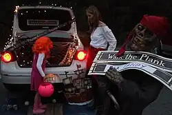 A parked white sedan is seen decorated with pirate-related Halloween decorations as a child in an orange wig takes candy from the trunk of the vehicle; a skeleton pirate statue holds the sign with the words "Walk the Plank FOR CANDY".