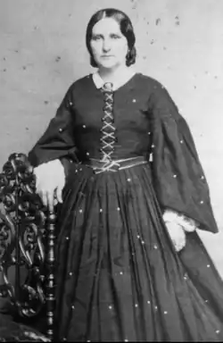 19th-century B&W portrait photo of a standing woman by a chair wearing a long, dark dress with a white collar, her hair parted and pulled back.