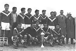 A group of men congregate around a silver trophy on the ground in this black-and-white photograph. Five men squat beside it, while ten stand behind. The five men at the front and six of those behind wear soccer attire; ten wear dark shirts marked with a white "V" extending from each shoulder to meet on the chest, with white shorts and hooped socks. The eleventh player, the goalkeeper, appears to be wearing all black. The four non-players wear street clothes; one wears a t-shirt and a greatcoat, while the other three wear suits and greatcoats.