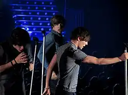 3 teenage boys singing into microphones on a stage.