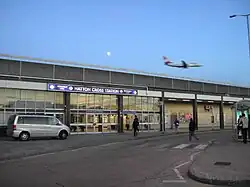 A dark grey building with a dark blue sign reading "HATTON CROSS STATION" in white letters all under a blue sky with an aeroplane flying through it