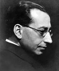 Monochrome portrait photo of a light-skinned, dark-haired man wearing glasses; he is captured in profile from the shoulder up, wearing dark clothing