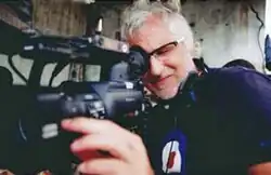 A handsome man with gray hair and glasses squints as he looks through a video camera's viewfinder while filming