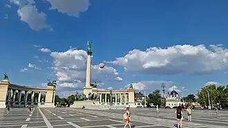 A wide-angle view of Heroes Square in Budapest showing the Millennium Monument, colonnades, and visitors in the open plaza under a clear sky.